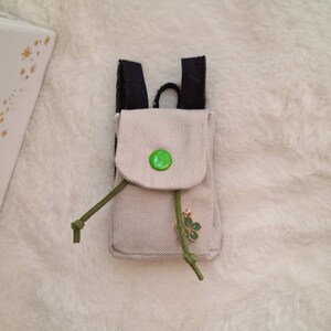 Könnte beinhalten: Ein kleiner beigefarbener Canvas-Rucksack mit schwarzen Trägern, einem grünen Knopf und einem grünen Kleeblatt-Anhänger.