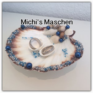 Könnte beinhalten: Ein weißer Muschelkasten für Ringe, verziert mit blauen und silbernen Perlen. Zwei silberne Eheringe liegen in der Muschel. Der Text "Michi's Maschen" steht über der Muschel.