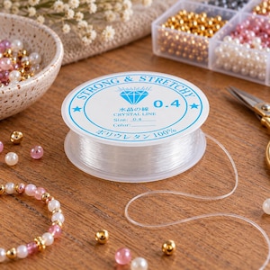 Könnte beinhalten: Eine Spule mit transparentem, elastischem Kristallfaden zur Schmuckherstellung. Das weiße Etikett zeigt ein blaues Diamantsymbol, den Text STRONG & STRETCHY, 0.4, CRYSTAL LINE sowie japanische Schriftzeichen für Kristallfaden und 100% Polyurethan.
