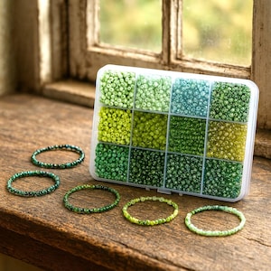 Può includere: Un organizer di plastica trasparente pieno di piccole perline rotonde in varie tonalità di verde e blu. Diversi braccialetti di perline in colori coordinati sono disposti su una superficie di legno, mostrando una gamma di tonalità verdi. I braccialetti hanno una circonferenza di circa 18 cm.
