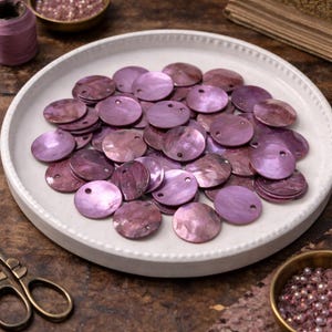 May include: A collection of iridescent, round, purple shell beads with small holes, displayed on a white ceramic plate. The beads vary in shades of purple and are arranged on a wooden surface, alongside a small bowl of beads and a spool of thread.