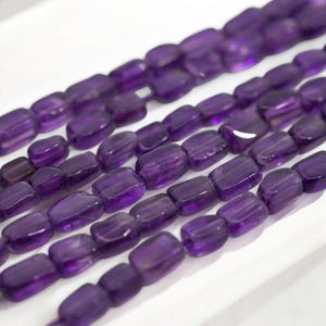 Könnte beinhalten: Eine Reihe von violetten Amethyst-Edelsteinkugeln, jede Kugel ist rechteckig und poliert, die Kugeln sind in Reihen auf einer weißen Oberfläche angeordnet.