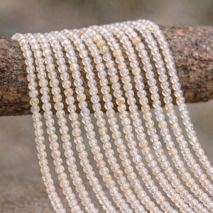 May include: Strands of small, round, light-colored beads, possibly gemstones, hang from a wooden branch. The beads have a translucent quality with subtle variations in color, ranging from clear to pale yellow.