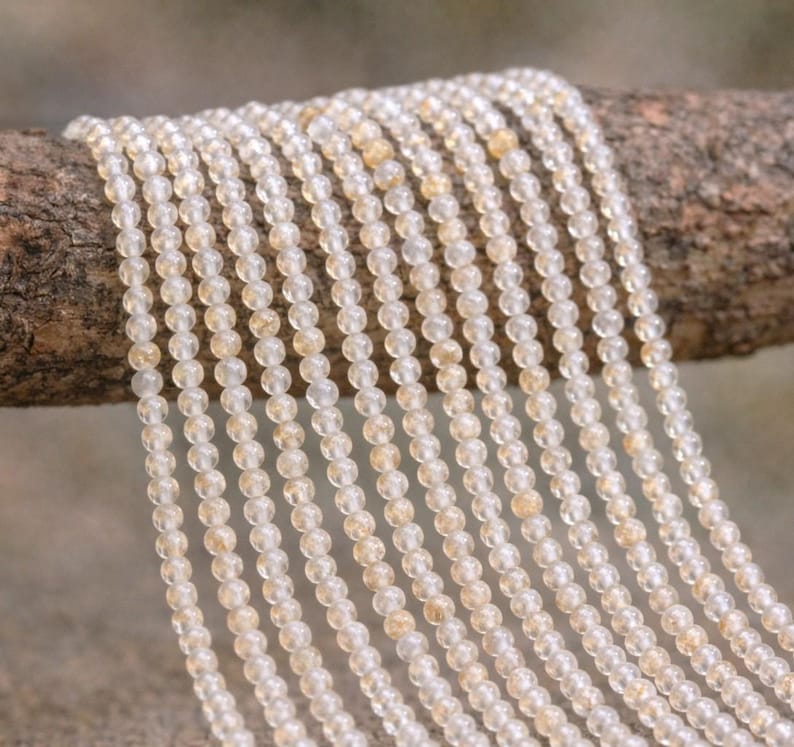 May include: Strands of small, round, light-colored beads, possibly gemstones, hang from a wooden branch. The beads have a translucent quality with subtle variations in color, ranging from clear to pale yellow.