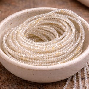May include: A collection of delicate, beaded necklaces in a cream-colored ceramic bowl. The necklaces are made of small, clear beads with subtle yellow accents. The bowl is off-white and sits on a textured, brown surface.