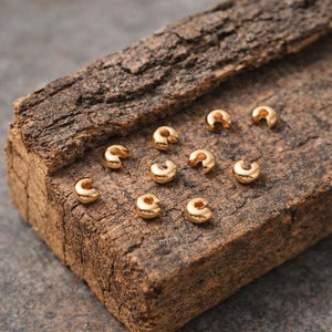 May include: A collection of small, gold-colored crimp beads, designed for jewelry making. The beads are C-shaped with a hole for stringing. They are displayed on a piece of rough, textured wood.