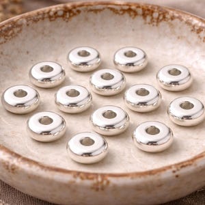 Könnte beinhalten: Eine Sammlung glänzender, silberfarbener, donut-förmiger Perlen mit einem zentralen Loch, präsentiert in einer hellbraunen Keramikschale. Diese Perlen werden wahrscheinlich für die Schmuckherstellung oder Bastelprojekte verwendet.