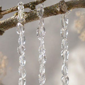 Op de afbeelding: Drie strengen heldere, gefacetteerde kristallen kralen hangen aan een natuurlijke tak. De kralen hebben een ovale en druppelvormige vorm en reflecteren licht. De achtergrond is zacht vervaagd.