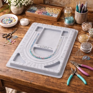 May include: A gray bead design board with a U-shaped channel for jewelry making. The board has measurement markings and holds beads, tools, and other supplies. A variety of beads in different colors are visible, along with jewelry-making tools.