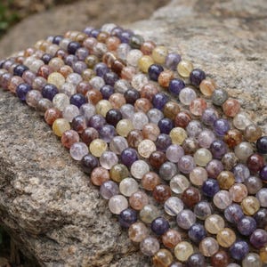 May include: A collection of gemstone beads in shades of purple, brown, yellow, and clear. The beads are spherical and strung together. The beads are displayed on a grey rock surface.