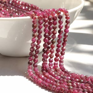 May include: Multiple strands of faceted, deep magenta-colored round beads are draped over the edge of a white bowl. Each bead features a reflective, multi-faceted surface that catches the light, creating a sparkling effect across the collection.