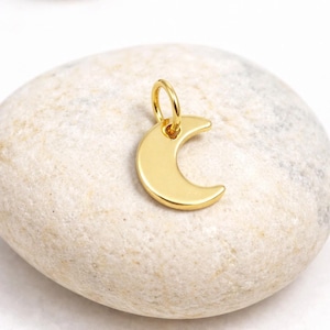 Op de afbeelding: Een goudkleurige maanvormige bedel met een kleine lus om aan een ketting of armband te bevestigen. De bedel is glad en glanzend en reflecteert licht. De maan staat tegen een neutrale achtergrond.