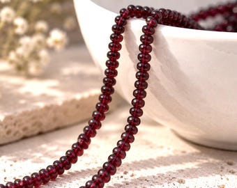 May include: A strand of small, round, deep red garnet beads. The beads are translucent and uniform in size, strung together in a continuous line. The strand is draped over the edge of a smooth, white ceramic bowl, showcasing the dark red color.