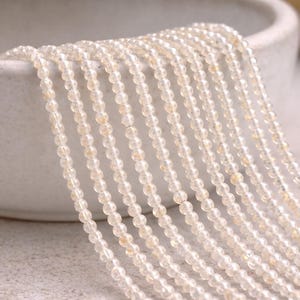 May include: Strands of small, round, light-colored beads cascade from a white ceramic bowl. The beads have a translucent quality with subtle variations in color, creating a delicate and elegant appearance. The beads are likely used for jewelry making.