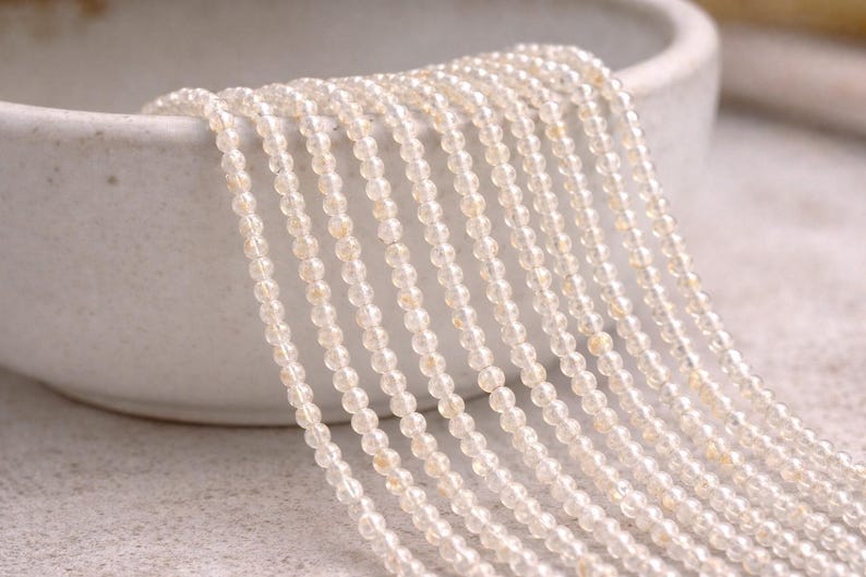 May include: Strands of small, round, light-colored beads cascade from a white ceramic bowl. The beads have a translucent quality with subtle variations in color, creating a delicate and elegant appearance. The beads are likely used for jewelry making.