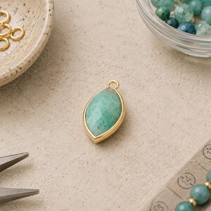 May include: A marquise-shaped pendant featuring a faceted green gemstone set in a gold-toned metal bezel with a small top loop. The pendant is surrounded by gold jump rings, a glass bowl of green beads, and metal jewelry pliers.