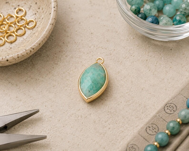 May include: A marquise-shaped pendant featuring a faceted green gemstone set in a gold-toned metal bezel with a small top loop. The pendant is surrounded by gold jump rings, a glass bowl of green beads, and metal jewelry pliers.
