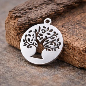 May include: A silver-colored pendant with a tree of life design. The pendant is circular with a cutout tree motif, featuring branches and leaves. It has a small loop at the top for a chain. The pendant is displayed on a textured wooden surface.
