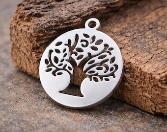 Puede incluir: Un colgante plateado con un diseño de árbol de la vida. El colgante circular presenta un motivo de árbol recortado, con ramas y hojas. Tiene un pequeño lazo en la parte superior para una cadena. El colgante se muestra sobre una superficie de madera texturizada.