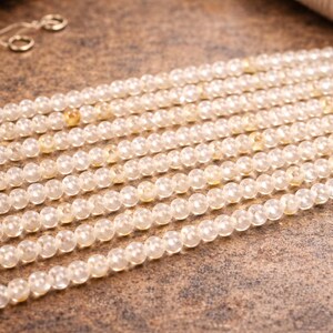 May include: Close-up of multiple strands of small, round, light yellow gemstone beads. The beads are strung together, creating a textured surface. The background is a mottled brown.