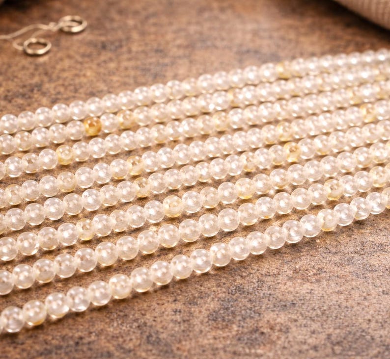 May include: Close-up of multiple strands of small, round, light yellow gemstone beads. The beads are strung together, creating a textured surface. The background is a mottled brown.