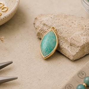May include: A marquise-shaped pendant featuring a faceted, teal-green gemstone set in a smooth gold-toned metal bezel. The pendant includes a small circular jump ring at the top for attachment, showcasing a polished finish and a pointed design.