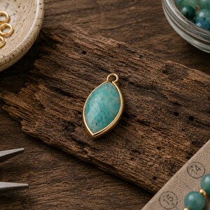 May include: A marquise-shaped pendant featuring a faceted teal amazonite stone set in a smooth gold-toned metal bezel. The pendant includes a small circular jump ring at the top for attachment, showcasing a polished finish and natural stone texture.