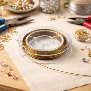 Pode incluir: Um carretel de fio dourado para miçangas é o ponto focal desta imagem, rodeado por materiais para fazer joias. Potes pequenos de contas, alicates e outros componentes estão dispostos sobre uma superfície clara, sugerindo um tema de artesanato ou joalheria.