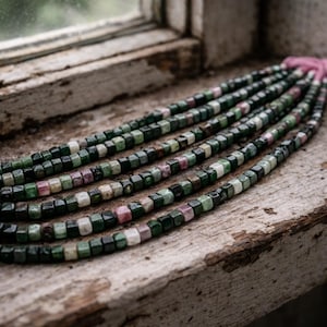 Könnte beinhalten: Mehrere Stränge aus Edelsteinperlen in Grün, Rosa und Weiß. Die kleinen, quadratischen Perlen sind aufgefädelt. Die Perlen liegen auf einer verwitterten Holzoberfläche in der Nähe eines Fensters.