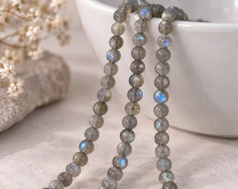 Puede incluir: Un hilo de cuentas redondas y pulidas de labradorita con una base de color gris y destellos iridiscentes en azul y dorado. Las cuentas están ensartadas en una sola línea, mostrando las variaciones naturales y propiedades de la piedra.