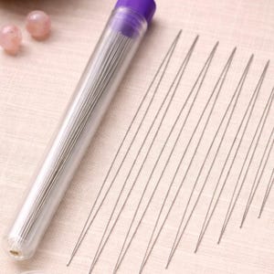 Può includere: Un tubo di plastica trasparente con un tappo viola pieno di sottili aghi per perline argentati. Diversi aghi sono sparsi su un tessuto rosa chiaro. Due perline rosa sono visibili sullo sfondo, suggerendo un tema artigianale.
