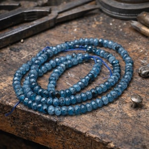 Op de afbeelding: Strengen gefacetteerde, blauwgroene edelsteen kralen zijn opgerold op een houten oppervlak. De kralen zijn geregen op een blauw koord. De achtergrond omvat metalen gereedschappen, wat wijst op een omgeving voor het maken van sieraden. De kralen hebben een glanzende, gepolijste uitstraling.