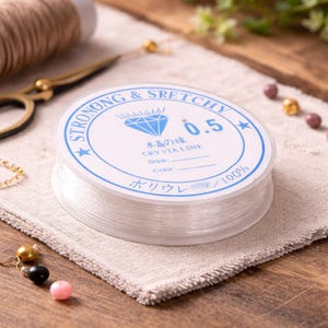 May include: A spool of clear, stretchy crystal line, labeled "STRONG & STRETCHY" with the size 0.5. The spool sits on a beige linen cloth, with gold scissors and beads in the background. The text "CRYSTAL LINE" is also visible.