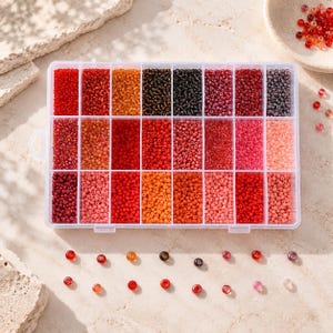 Op de afbeelding: Een rechthoekige doorzichtige plastic opbergdoos met vierentwintig individuele vakjes gevuld met kleine glazen kraaltjes in diverse tinten rood, oranje, roze, bruin en zwart. Enkele losse kraaltjes liggen verspreid buiten de doos.