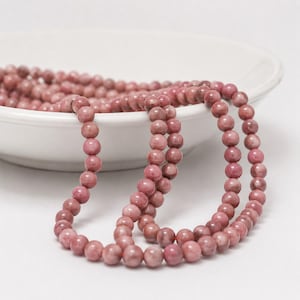 May include: A strand of round, dusty rose-colored beads with subtle natural veining and variations in tone. The beads are strung together in a continuous loop, with a portion of the strand resting inside a white bowl and the rest draped forward.