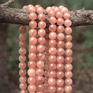 Könnte beinhalten: Stränge aus pfirsichfarbenen, runden Edelsteinperlen hängen an einem Ast. Die Perlen haben eine glatte, polierte Oberfläche und einen subtilen Schimmer. Der Hintergrund ist ein verschwommenes Grün, was auf eine natürliche Umgebung hindeutet. Geeignet für die Schmuckherstellung.