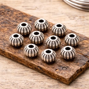 May include: Close-up of ten silver-colored, ribbed beads with large holes, arranged on a rustic wooden surface. The beads have a detailed, textured design and are likely used for jewelry making or crafting. The beads are round and have a metallic sheen.