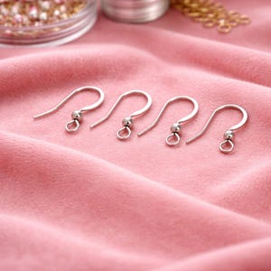 May include: Four silver-colored earring hooks with a ball and loop design. The hooks are arranged on a pink fabric surface, with other jewelry-making supplies visible in the background. These components are for DIY jewelry making.