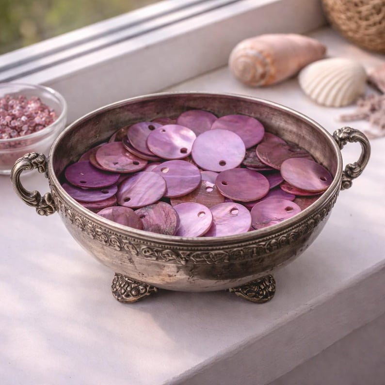 Op de afbeelding: Een zilveren kom gevuld met ronde, iriserende paarse pailletten. De kom heeft sierlijke handvatten en staat op kleine voetjes. Een kleine glazen container met roze korrels is links zichtbaar, met schelpen op de achtergrond.
