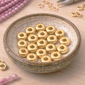 Könnte beinhalten: Nahaufnahme von goldfarbenen, donut-förmigen Perlen in einer kleinen, gesprenkelten Keramikschale. Im Hintergrund sind weitere goldfarbene Schmuckzubehörteile und rosa Perlen zu sehen. Die Perlen haben einen Durchmesser von etwa 0,6 cm.