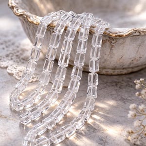 Puede incluir: Varias hebras de cuentas rectangulares transparentes caen de un cuenco decorativo blanquecino. Las cuentas transparentes reflejan la luz, lo que sugiere que están hechas de vidrio o cristal. El fondo es una superficie neutra y texturizada con iluminación suave.