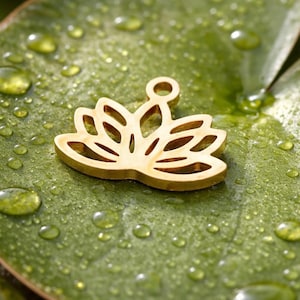 Op de afbeelding: Een goudkleurige lotusbloemhanger met een lus voor een ketting. De hanger ligt op een groen blad bedekt met waterdruppels. Het lotusbloemontwerp heeft meerdere uitgesneden secties.