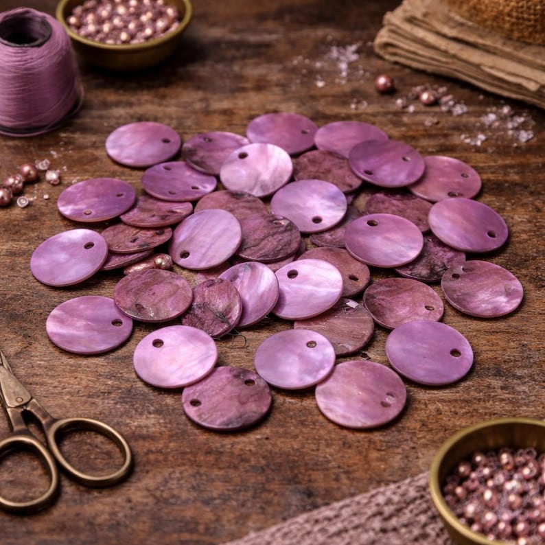 Op de afbeelding: Een verzameling platte, ronde, paarse schelpkralen met een gat in het midden, verspreid op een houten oppervlak. De kralen vari&euml;ren in tinten paars. Andere knutselbenodigdheden, waaronder een klos garen, kleine bakjes met kralen en een schaar, zijn ook zichtbaar.