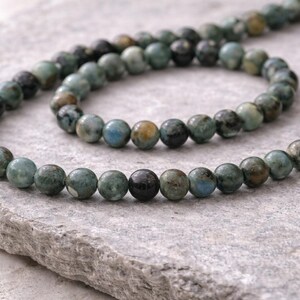 May include: A string of round gemstone beads in shades of teal, blue, and brown. The beads are displayed on a grey, textured surface. The beads are approximately 8mm in diameter, ideal for jewellery making.