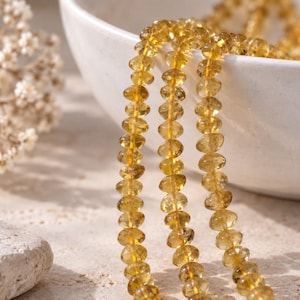 May include: A strand of faceted, translucent yellow citrine beads arranged in three vertical rows. Each bead features a circular, flattened shape with a polished finish that catches the light, creating a bright, golden appearance throughout the strand.
