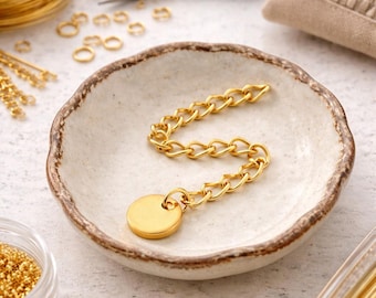 May include: A gold-colored chain with a circular pendant rests in a small, off-white ceramic dish. The chain is made of interlocking links. Jewelry-making supplies, including wire, jump rings, and tools, are in the background.