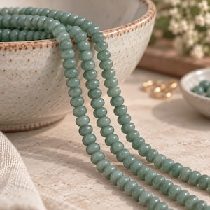May include: Three strands of smooth, round, sage green beads are draped over the edge of a speckled ceramic bowl. The beads have a uniform size and a soft, matte finish, creating a continuous line of color that contrasts with the bowl.