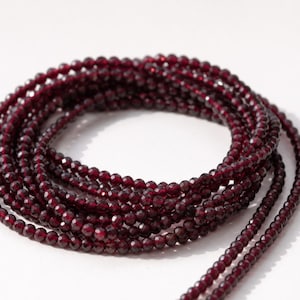 May include: A coiled strand of small, faceted, deep red beads. The beads are strung together, forming a circular shape. The beads have a glossy finish and are displayed against a white background. The beads appear to be gemstones.