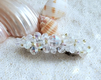 May include: A clear crystal hair clip with iridescent beads and gold accents. The clip is on a white background with seashells.