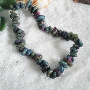 May include: A beaded bracelet composed of irregular, tumbled gemstone chips in shades of deep forest green, teal, and vibrant magenta. The stones are strung together in a continuous loop, showcasing natural variations in color, texture, and shape.